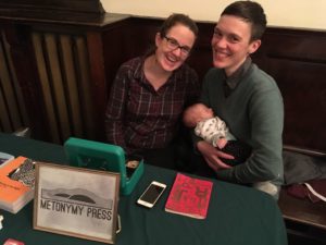 Two adults lean cheeks towards each other, smiling, and one of them holds a small sleeping baby. A framed Metonymy Press sign, a cashbox, a box, a notebook, and a phone are on the table in front of them.