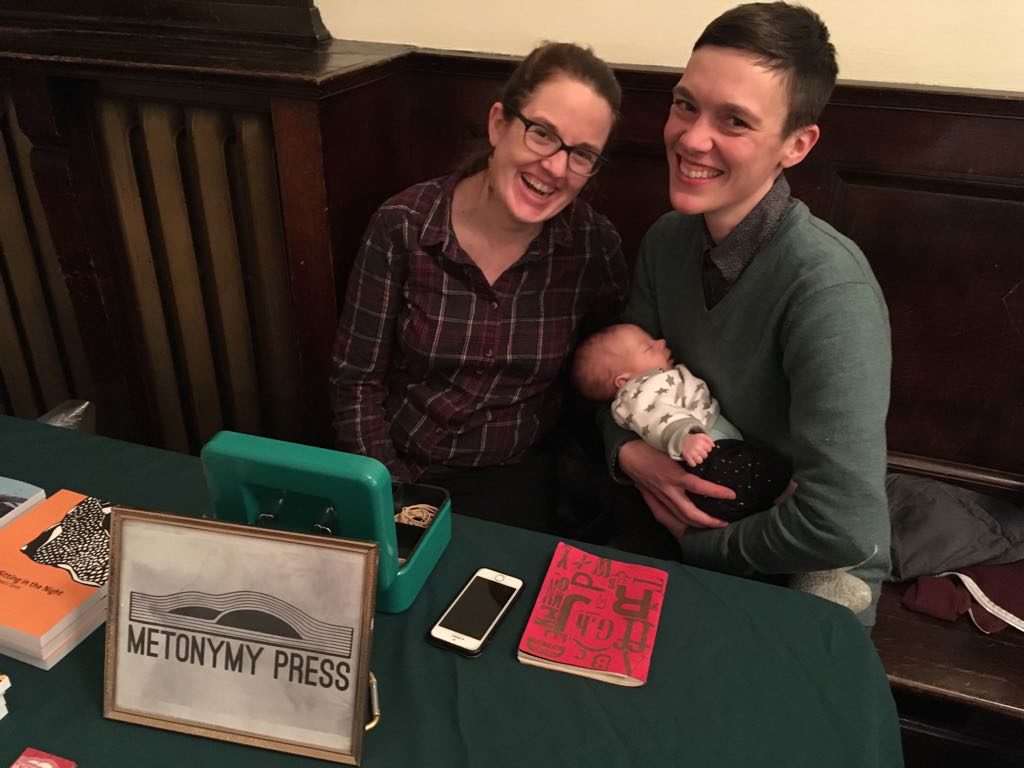 Two adults lean cheeks towards each other, smiling, and one of them holds a small sleeping baby. A framed Metonymy Press sign, a cashbox, a box, a notebook, and a phone are on the table in front of them.