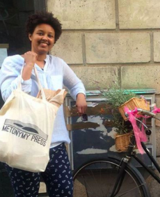 Friend of the publishers leans against a wall near a bicycle, smiling, holding up a full Metonymy tote bag.