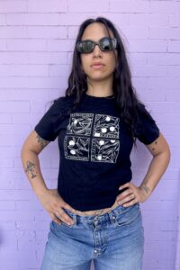 Photograph of model Kristeen Lynch wearing a black Metonymy t-shirt designed by Lee Lai against a purple brick background.